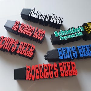 Könnte beinhalten: Sechs schwarze Zapfhähne mit roter, blauer, weißer und grüner Beschriftung. Die Griffe sind mit "Aaron's Beer", "El Torro Loco", "John's Beer", "Schneider's Bayrisch Hall", "Ben's Beer" und "Robert's Beer" beschriftet.