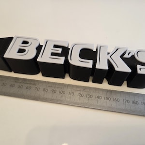 Könnte beinhalten: Ein 3D-gedrucktes Schild mit dem Schriftzug "BECK'S" in Schwarz und Weiß. Die Buchstaben sind dick und blockartig, mit weißen Umrissen. Ein Metalllineal liegt darunter und misst in Zentimetern.