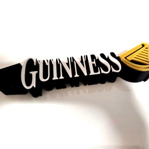 Könnte beinhalten: Ein schwarz-weißes Guinness-Schild mit goldenem Akzent. Das Wort "GUINNESS" ist in weißen, erhabenen Buchstaben geschrieben. Das Schild hat eine schwarze Basis und ein gold-schwarzes Emblem auf der rechten Seite.