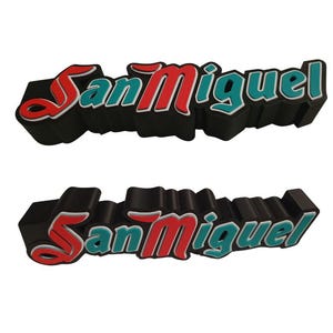 Könnte beinhalten: Ein schwarzes 3D-Schild mit dem Text "San Miguel" in roten und türkisfarbenen Buchstaben.