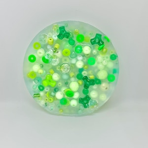 May include: A clear round disc filled with green and white beads, including small round beads, larger round beads, and star-shaped beads.