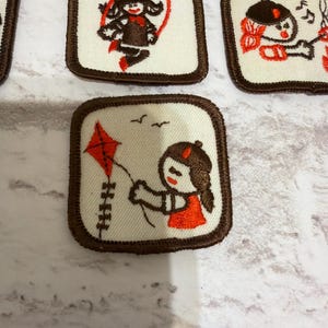 May include: A selection of square patches with brown edging and embroidered designs. The patches depict cartoon girls engaged in activities such as skipping, playing music, and flying a kite. The colour scheme includes white, brown, and orange.