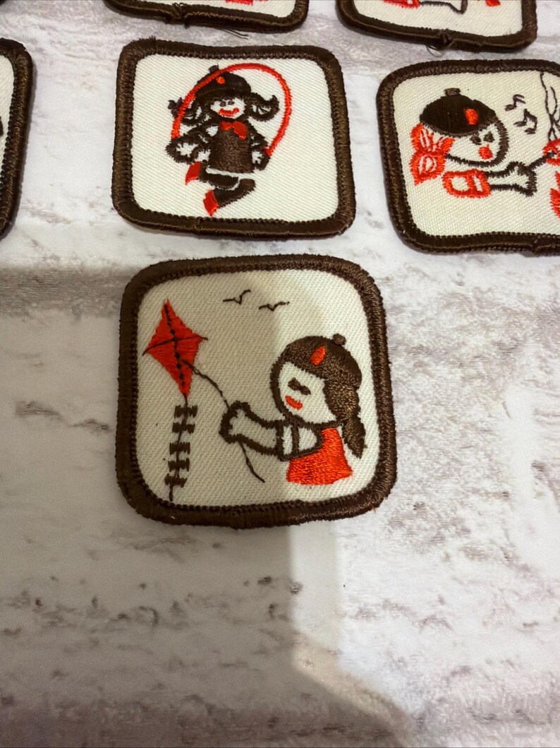 May include: A selection of square patches with brown edging and embroidered designs. The patches depict cartoon girls engaged in activities such as skipping, playing music, and flying a kite. The colour scheme includes white, brown, and orange.