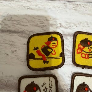 May include: Several embroidered patches with cartoon figures. The patches have a yellow or white background and brown borders. One patch shows a figure jumping over a line, while others depict different scenes.