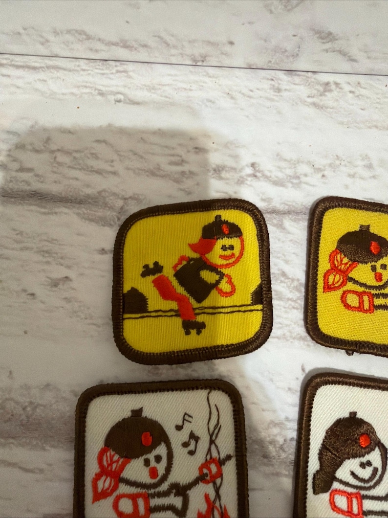 May include: Several embroidered patches with cartoon figures. The patches have a yellow or white background and brown borders. One patch shows a figure jumping over a line, while others depict different scenes.