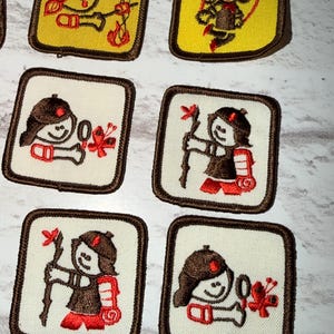 May include: A selection of embroidered patches, each showcasing a cartoon figure in outdoor pursuits. The patches have either a white or yellow backdrop, framed by brown borders. The character, wearing a brown hat and red details, is shown engaged in activities such as playing music, skipping, and hiking.