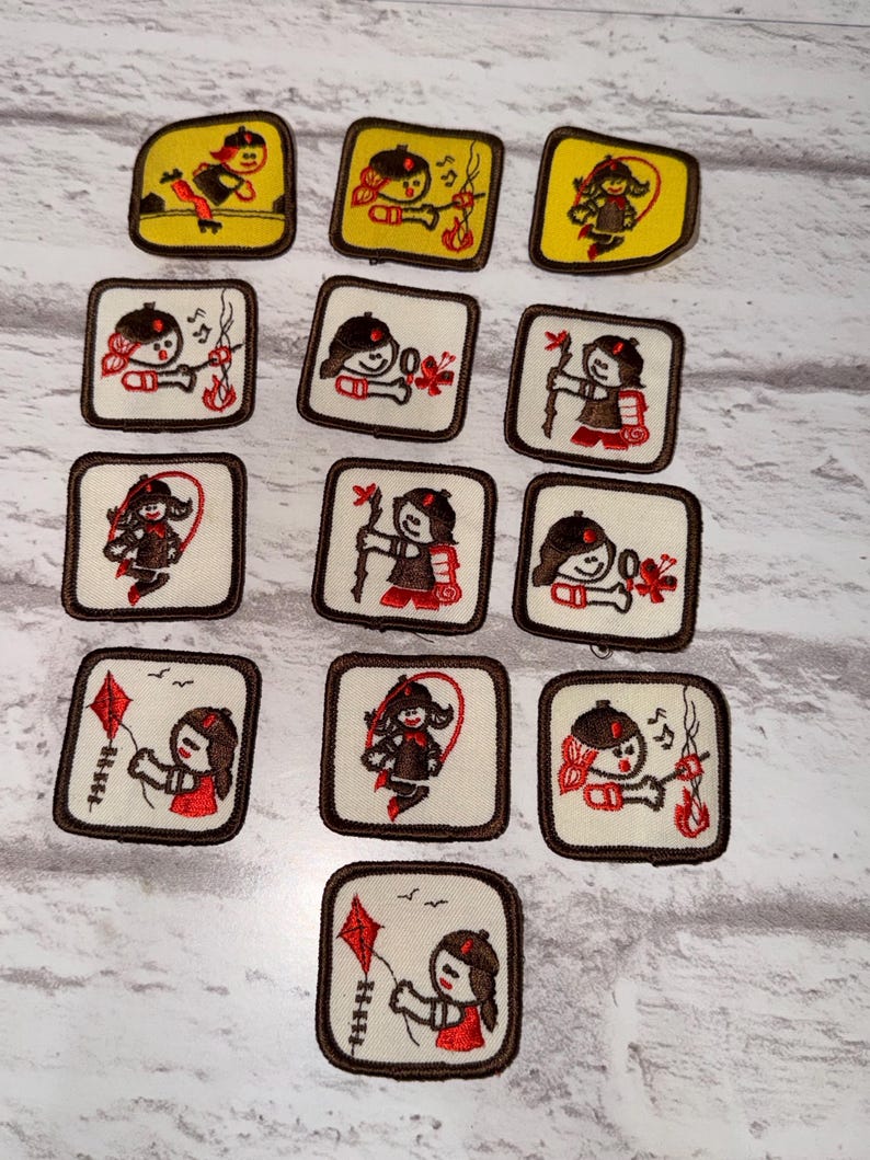 May include: A selection of embroidered patches with a brown border, showcasing a cartoon girl engaged in outdoor pursuits. The patches depict activities such as playing a musical instrument, archery, and flying a kite. The backgrounds are either yellow or white.