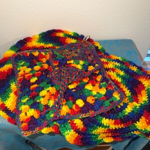 May include: A colorful, handmade crochet set featuring a blanket and a square pouch. The blanket has a circular shape with a rainbow pattern. The pouch is square with a similar rainbow design. The set is made with vibrant red, orange, yellow, green, blue, and purple yarn.