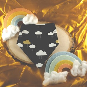 May include: A dark gray bandana with a white cloud and rain pattern, and gold clouds. The bandana is displayed on a wooden slice with felt rainbows and cotton clouds. The background is a golden fabric.