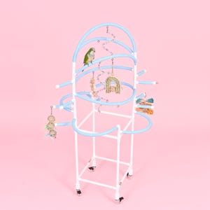 May include: A white and light blue bird play stand with a green parrot perched on a curved perch. The stand has multiple levels with hanging toys and a pink background. The stand is on wheels.