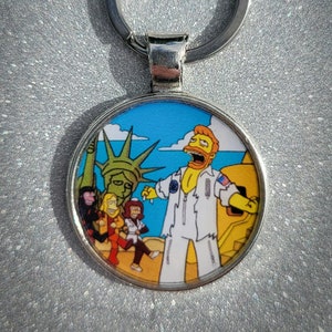 May include: A silver keychain with a round, colorful image of a cartoon character with a yellow beard and a white jumpsuit standing in front of a statue of liberty. The character is holding a yellow object in their right hand and is smiling with their mouth open. There are three other cartoon characters in the background.