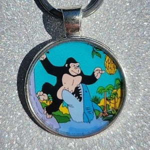 May include: A silver keychain with a round, colorful image of a gorilla riding a shark. The gorilla is wearing a blue shirt and has a banana in its hand. The background is a blue ocean with palm trees.