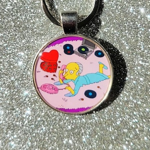 May include: A round keychain featuring a cartoon character on a pink background. The character is yellow with blue clothing and is talking on a pink phone. There are hearts, records, and a record player in the background.