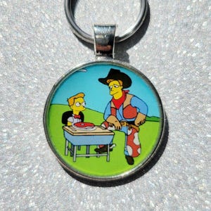 May include: A round metal keychain with a cartoon image of two characters. The character on the left is a yellow-skinned boy with brown hair wearing a blue shirt and a red bow tie. The character on the right is a yellow-skinned man with brown hair wearing a cowboy hat, a red vest, and blue jeans with white spots. The characters are standing in front of a blue and green background.