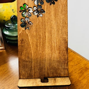 May include: A wooden phone stand with a dark brown stain. The stand has a cutout design of five hibiscus flowers in the top right corner.