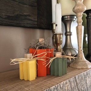 Pode incluir: Um conjunto de três abóboras de madeira pintadas de amarelo, laranja e verde. As abóboras são decoradas com palha e estão sentadas em uma prateleira de madeira com castiçais no fundo.
