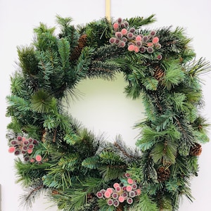 Könnte beinhalten: Ein grüner Weihnachtskranz mit Tannennadeln und braunen Tannenzapfen, verziert mit rosa Beeren. Der Kranz hängt an einer weißen Wand mit einem goldenen Stern oben.