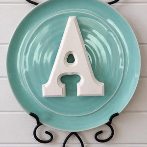 Op de afbeelding: Een witte houten letter "A" is gecentreerd op een turquoise bord, gemonteerd op een zwarte metalen bordenhouder. Het bord heeft een geribbeld ontwerp. De achtergrond is een witte houten muur.