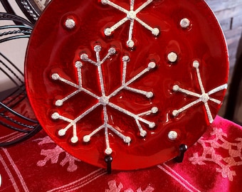 May include: A round, red decorative plate with silver snowflake designs. The plate is displayed on a black stand, set against a red patterned fabric. The plate is a festive holiday decoration.