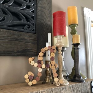 Op de afbeelding: Een houten plank met een kurken letter A, drie kandelaars en drie kaarsen. De kandelaars zijn gemaakt van metaal en hout. De kaarsen zijn rood, geel en wit.