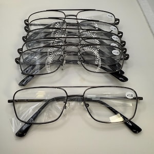 May include: A stack of black metal-framed reading glasses with clear lenses. Each pair has a +1.00 strength marking on the lens. The glasses are rectangular and have a classic design.