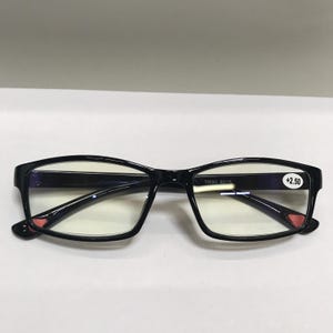 May include: Black rectangular eyeglasses with clear lenses. The frames have a glossy finish and small red accents near the hinges. The right lens has a sticker with the text "+2.50".