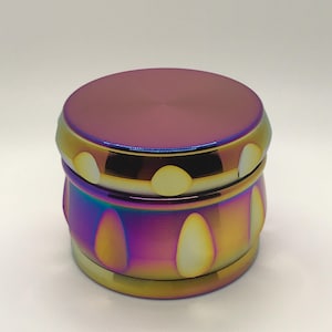 May include: A metallic grinder with a color-shifting finish, transitioning from purple to gold and blue. The top and base are a deep purple, while the body features a series of teardrop-shaped cutouts. The grinder is designed with a cylindrical shape.
