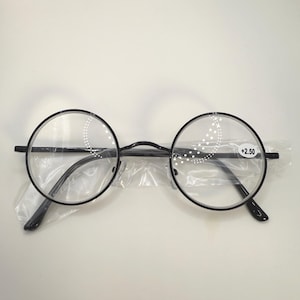 May include: Round black eyeglasses with clear lenses. The glasses have a thin black frame and are in a clear plastic sleeve. The right lens has the text "+2.50".