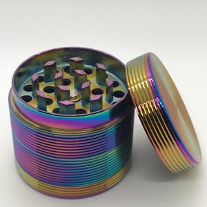 May include: A multi-colored, rainbow-hued grinder with a screw-on lid. The grinder is cylindrical with a ridged exterior and a top section with sharp, square teeth. The lid is off to the side, revealing the interior grinding components.
