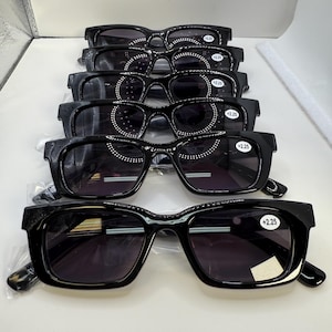 May include: A stack of six black sunglasses with dark lenses. Each pair has a thick, rectangular frame. The lenses have a +2.25 strength sticker. The sunglasses are displayed on a white surface.