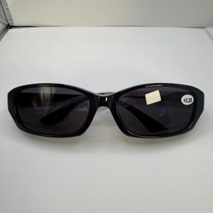 May include: Black sunglasses with dark lenses. The glasses have a rectangular shape with rounded edges. The right lens has a sticker with the text "+2.25". The sunglasses are on a white surface.
