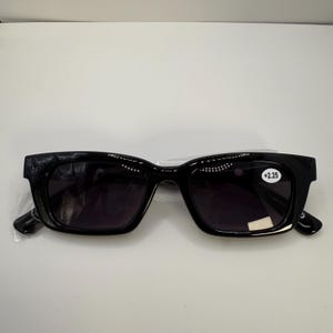 May include: Black rectangular sunglasses with dark lenses. The glasses have a thick frame and are marked with a +2.25 magnification. The sunglasses are in a clear plastic wrapper.