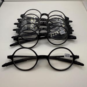 May include: A stack of black, round-framed reading glasses with clear lenses. The glasses have a folding design and are displayed on a white surface. Each pair has a small sticker with text.