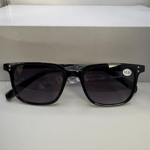 May include: Black square-framed sunglasses with dark lenses. The glasses have a +2.25 strength marking on the right lens. The frames are thick and have small silver accents on the temples. The sunglasses are in a clear plastic sleeve.