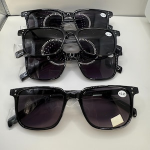 May include: Four pairs of black square-framed sunglasses with dark lenses are stacked. Each pair has a small white sticker with the text '+2.25' on the upper right corner of the frame. The sunglasses are new and still in their protective plastic.