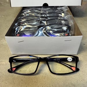 May include: A box of black-framed reading glasses with clear lenses. One pair of glasses is displayed in front of the box. The glasses have a rectangular shape and red accents on the temples. The text "+2.00" is visible on the lens.