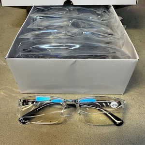 May include: A box of clear-framed reading glasses with black temples. One pair of glasses is in the foreground, displaying a +3.00 magnification. The box contains multiple pairs of glasses, each individually packaged in clear plastic.