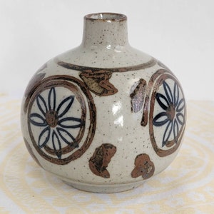 May include: A small, round, white ceramic vase with brown and black floral designs. The vase has a speckled glaze and a narrow neck.