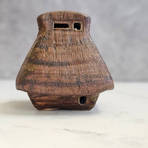 May include: A small, brown ceramic house with a textured surface and two small windows. The house has a rounded roof and a small opening at the bottom.