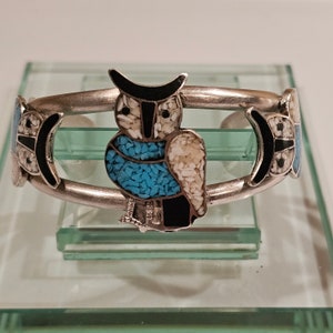 May include: Silver-toned cuff bracelet featuring an owl design. The owl is adorned with turquoise, black, and white mosaic inlays. The bracelet has a curved design with decorative accents on each side.