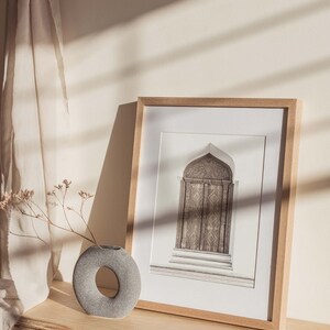 May include: A framed black and white photograph of an ornate wooden door with an arched top. The frame is made of light wood and is sitting on a light wood shelf with a gray stone vase and dried flowers.