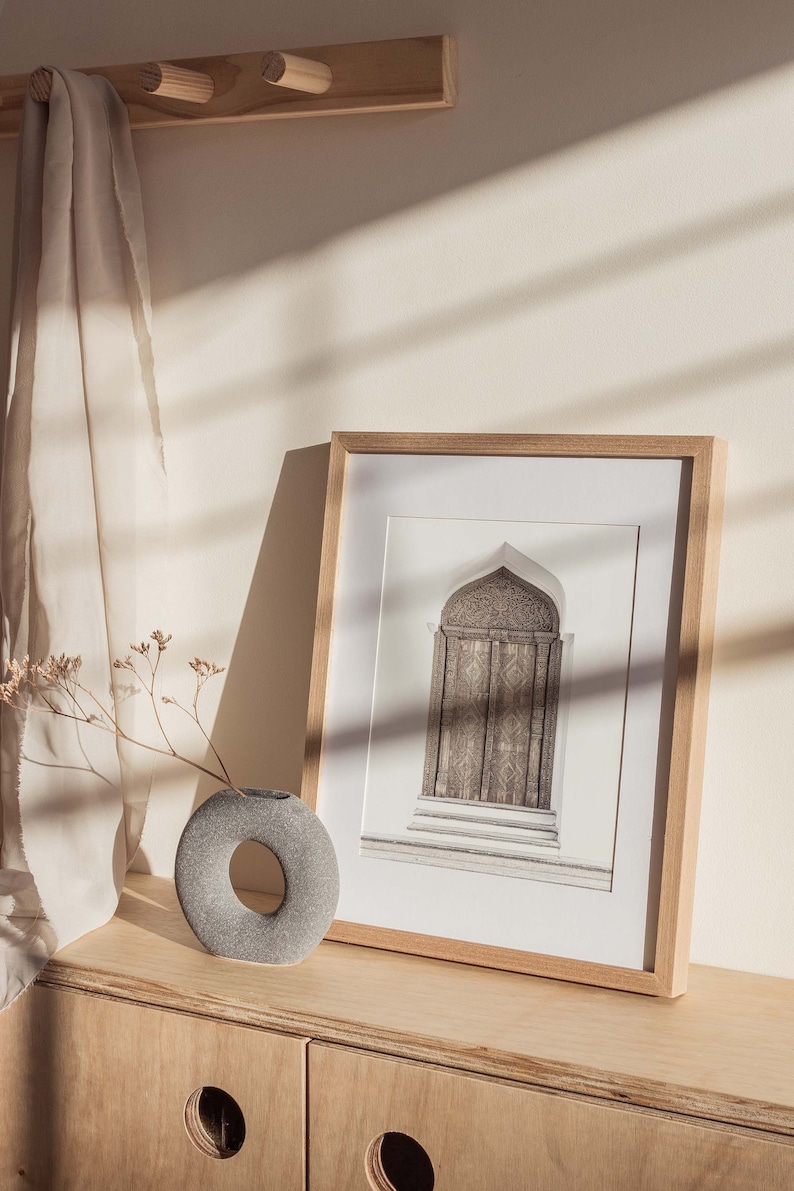 May include: A framed black and white photograph of an ornate wooden door with an arched top. The frame is made of light wood and is sitting on a light wood shelf with a gray stone vase and dried flowers.