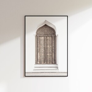 May include: A framed photograph of a carved wooden door with an arched top. The door is brown and has intricate details. The door is set against a white wall.