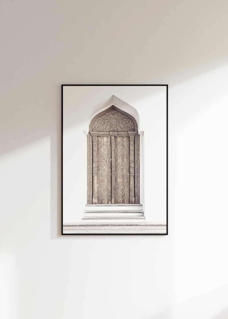 May include: A framed photograph of a carved wooden door with an arched top. The door is brown and has intricate details. The door is set against a white wall.