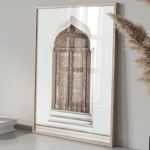 May include: A framed print of a photograph of a carved wooden door with intricate details. The door is arched and has a light brown color. The background is a white wall.