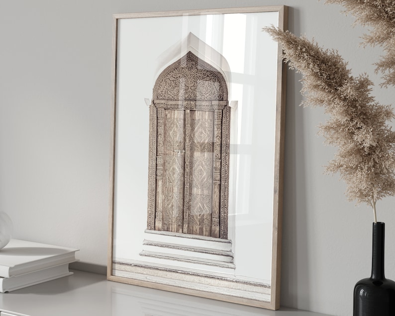 May include: A framed print of a photograph of a carved wooden door with intricate details. The door is arched and has a light brown color. The background is a white wall.