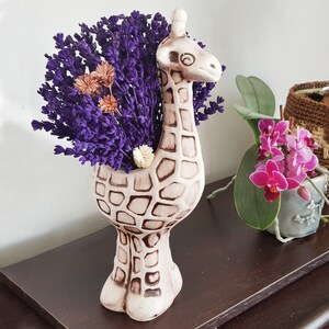 May include: A ceramic giraffe planter with a brown and white pattern. The planter is filled with purple lavender flowers.