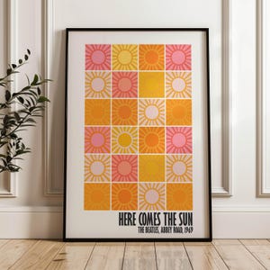Peut inclure: Impression d'art encadr&eacute;e avec une grille de soleils roses, jaunes et oranges. Le texte "HERE COMES THE SUN" et "THE BEATLES, ABBEY ROAD, 1969" est imprim&eacute; en noir. L'&oelig;uvre est dans un cadre noir. Le tout est pos&eacute; sur un sol en bois.