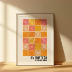 Peut inclure: Impression d'art encadr&eacute;e avec une grille d'illustrations de soleil dans des tons de rose, orange et jaune. Le texte "HERE COMES THE SUN" est affich&eacute; en dessous, avec "THE BEATLES, ABBEY ROAD, 1969". L'&oelig;uvre est dans un cadre blanc.