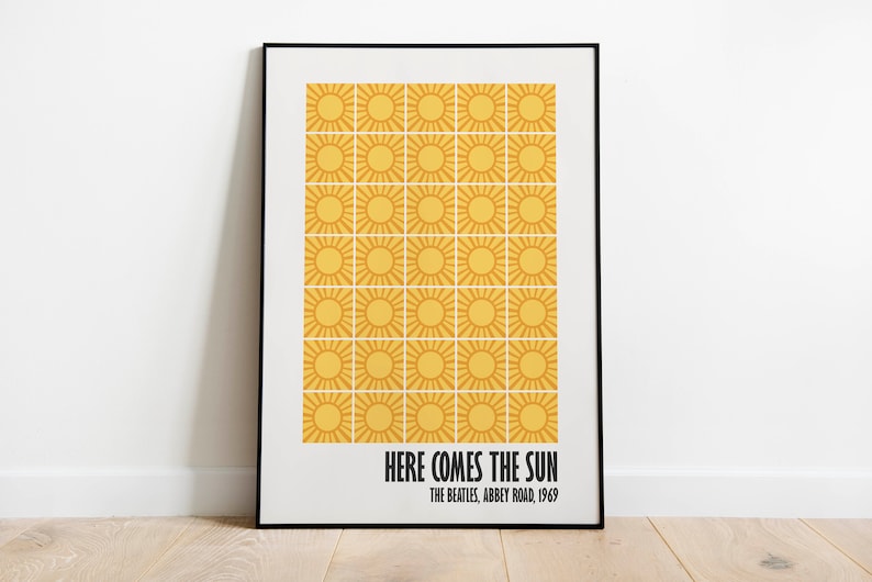 Peut inclure: Une affiche encadr&eacute;e avec une grille de soleils jaunes sur fond blanc. Le texte "HERE COMES THE SUN" et "THE BEATLES, ABBEY ROAD, 1969" est imprim&eacute; en noir. Le cadre est noir, et l'affiche est pos&eacute;e sur un sol en bois.